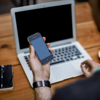 Ein Mann hält ein Smartphone vor seinem Arbeitsplatz mit Notizbuch und Laptop