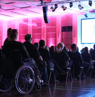 Bei der Auftaktveranstaltung des 16. Jugendhilfetags sitzt eine Rollstuhlfahrerin neben den anderen Teilnehmenden im Mittelgang mit Blick zur Bühne