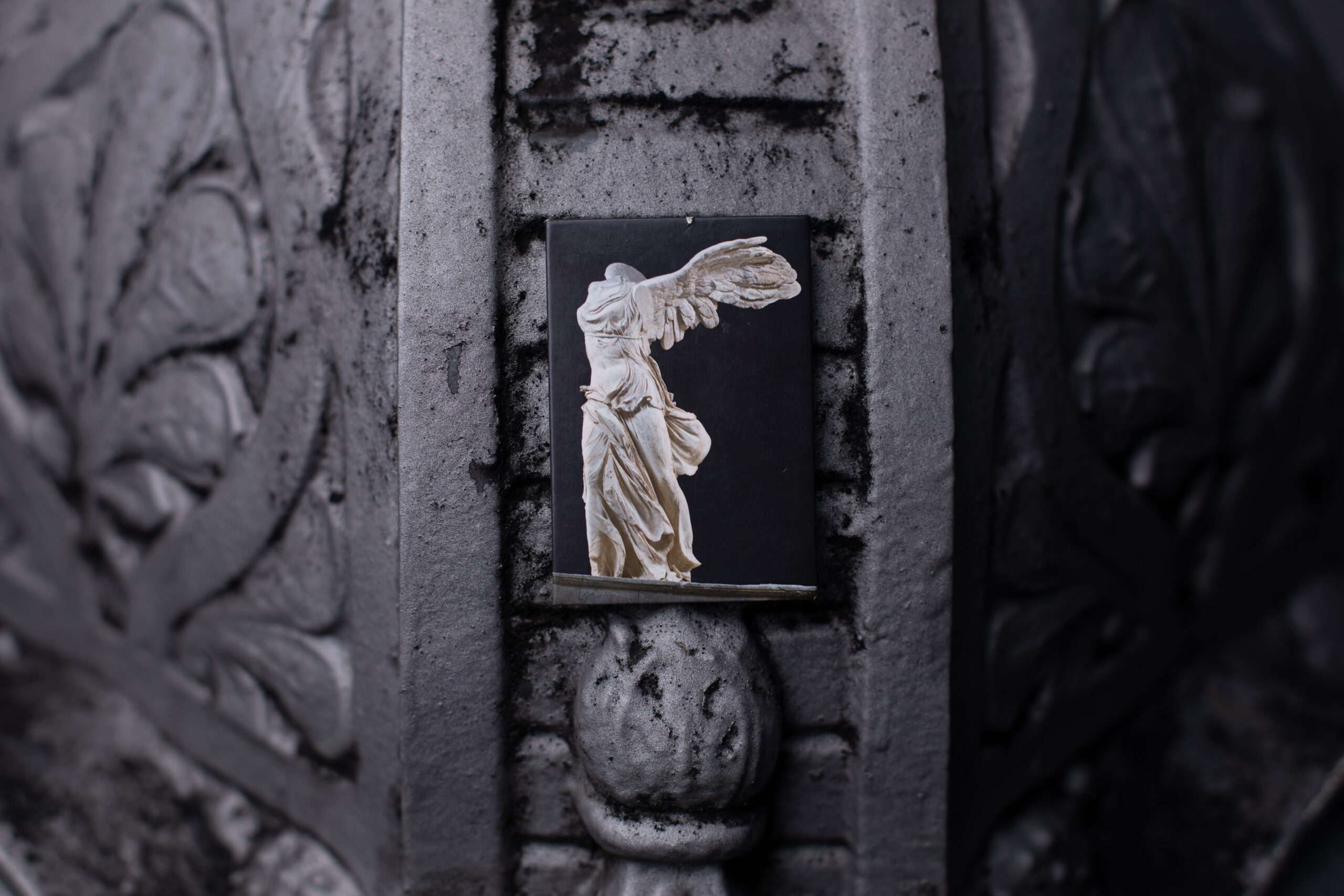 Nahaufnahme. An eine graue Laterne lehnt der Magnet mit der Siegesgöttin Nike. Ein rechteckiger Magnet mit schwarzem Hintergrund. Davor die weiße Skulptur, der Arme und Kopf fehlen. Sie befindet sich in einer Landeposition, ist mit offenen Flügeln leicht nach vorne übergebeugt. Die Flügel sind dadurch mit Luft erfüllt und nach hinten gedrückt, ähnlich wie das dünne flatternde Gewand, das an den Bauch gepresst ist. Ihr Bauch wirkt auf diese Weise entblößt. Sie verlagert ihr Gewicht leicht nach vorne auf das rechte Bein, ist aber insgesamt aufrecht.