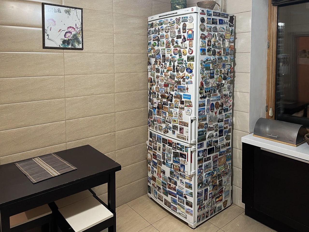 Ein Bild von Michaels Kühlschrank in Kiew. Der Kühlschrank ist weiß und besteht aus einem Kühl- und einem Gefrierfach, die übereinander liegen. Der Kühlschrank ist etwa zwei Meter groß und steht in der Ecke der Küche. Die linke Seite steht zur Wand. Die Front und die rechte Seite sind komplett mit Magneten bedeckt. Rechts neben dem Kühlschrank ist ein Fenster. Darunter ein schwarzer Schrank. Gegenüber dem Kühlschrank ist ein schwarzer Tisch, über dem ein Bild hängt. Die Küche ist beige gefliest.