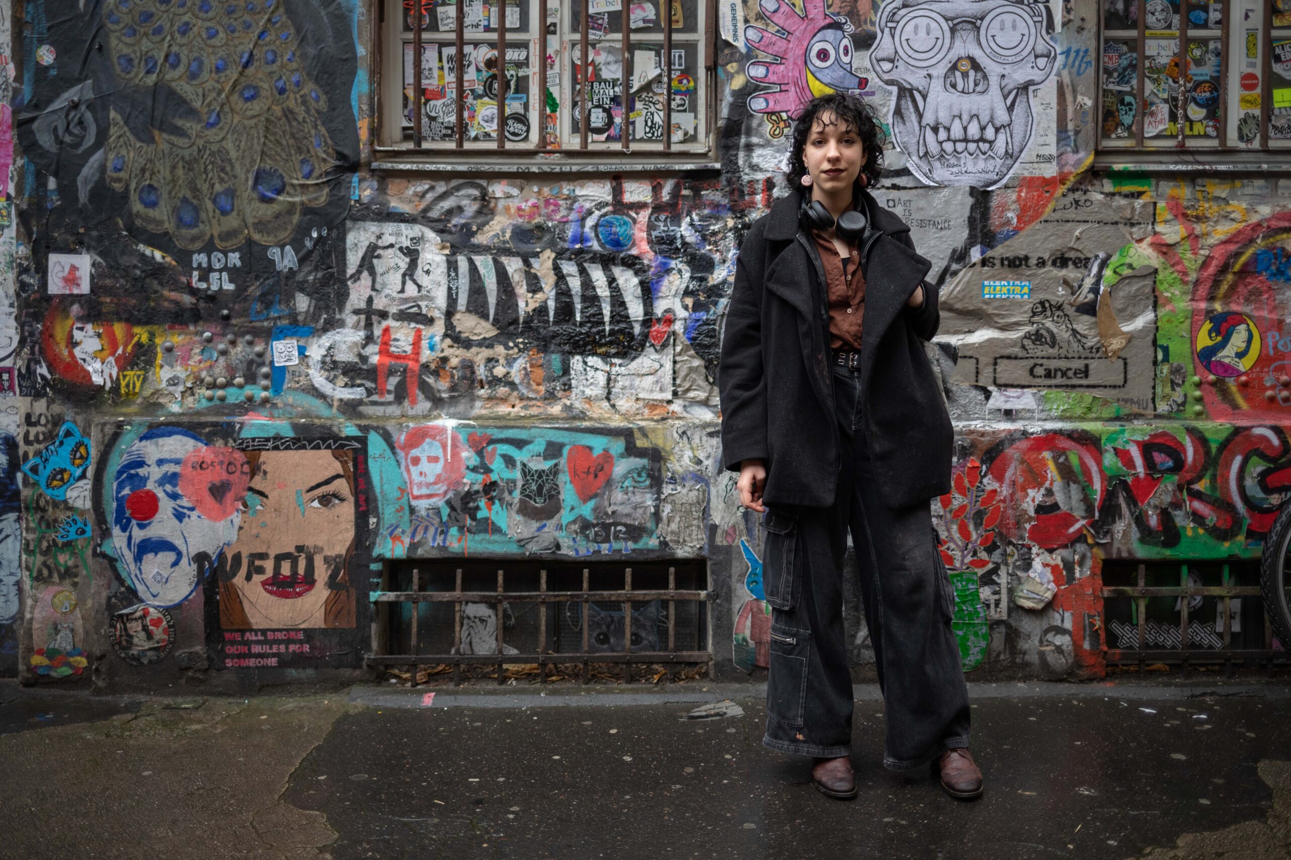 Auf dem Bild sieht man Nox vor einer grauen Betonwand stehen. Die Betonwand ist bemalt mit buntem Graffiti und beklebt mit einigen Stickern.Nox steht rechts im Bild, trägt eine schwarze Hose und einen schwarzen, langen Mantel. Um deren Hals hängen schwarze Kopfhörer. Nox hat schulterlange, lockige, schwarze Haare. Der Mantel ist auf und man sieht darunter eine braune Bluse.